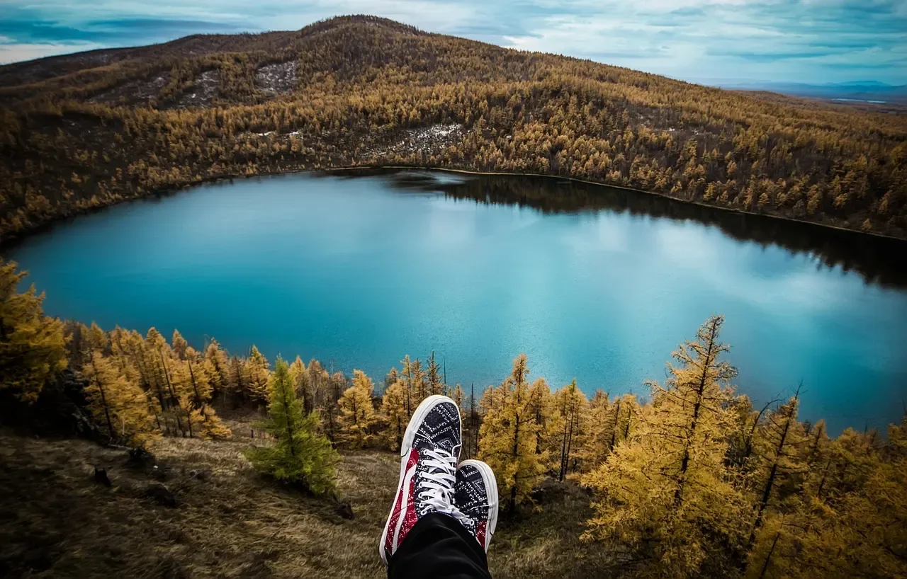 Füße in Sneakern auf einer Aussichtskante über einem blauen See und herbstlichem Wald.